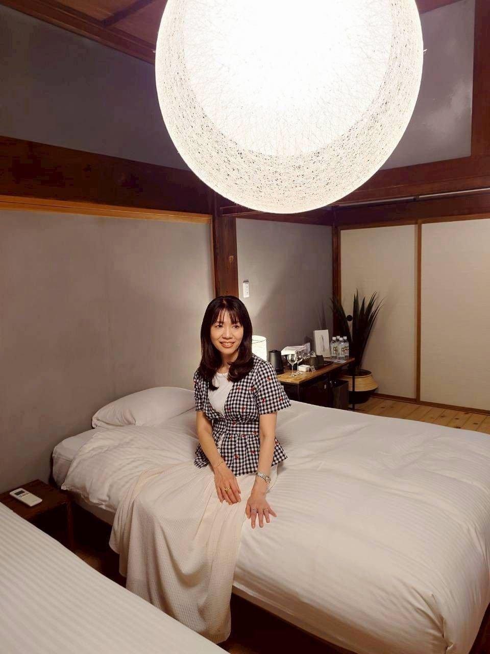 白を基調とした和風の部屋で、ベッドに座る女性。ZARAのギンガムチェリー刺繍トップスに白いスカートを着用。天井には大きな球体のペンダントライトが吊り下げられている。