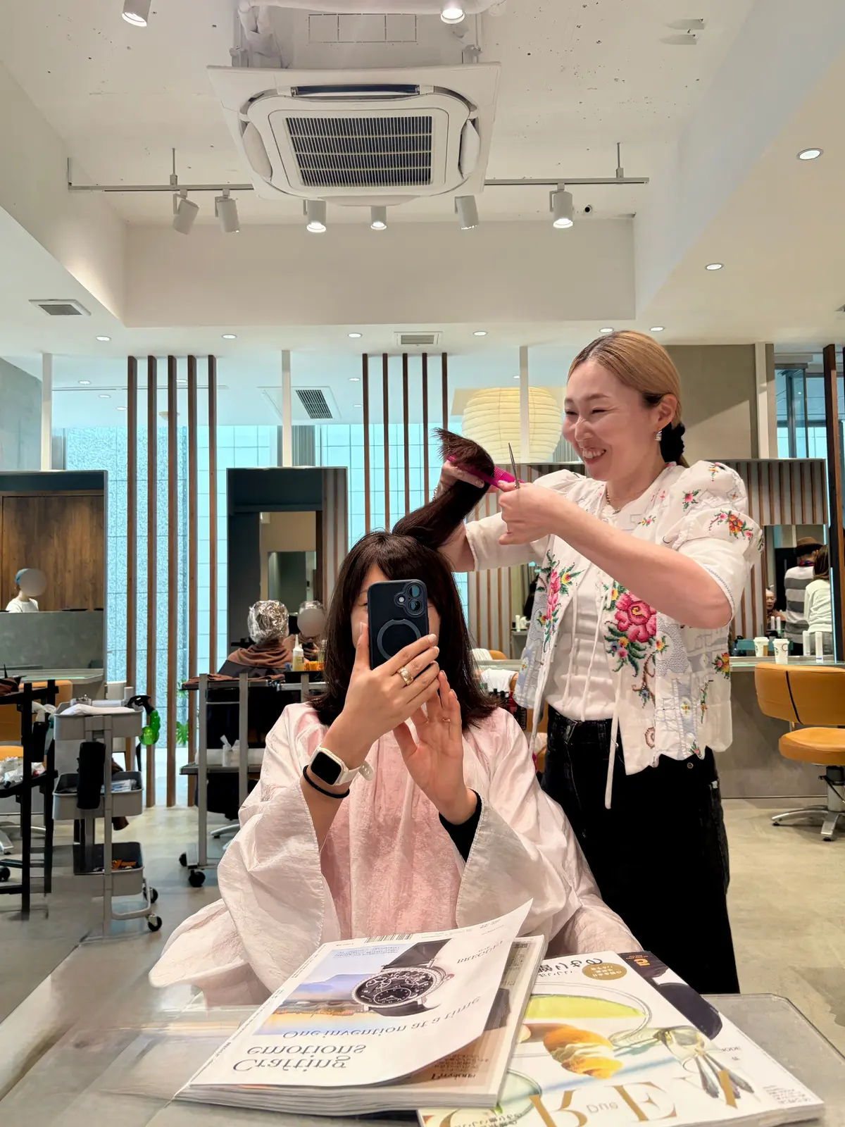 美容室で、スタイリストが女性客の髪をカットしている様子。女性客はスマートフォンで自撮りをしながら、カットされた髪をピンクのクリップで留めている。背景には他の客やスタッフが見える。