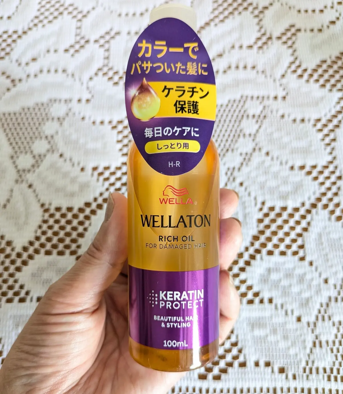 手に握られた、ウェラ(WELLA)の「ウェラトーン リッチオイル」のボトル。ピンクと水色のラベルに日本語で「ケラチン保護」の文字。容量は100ml。
