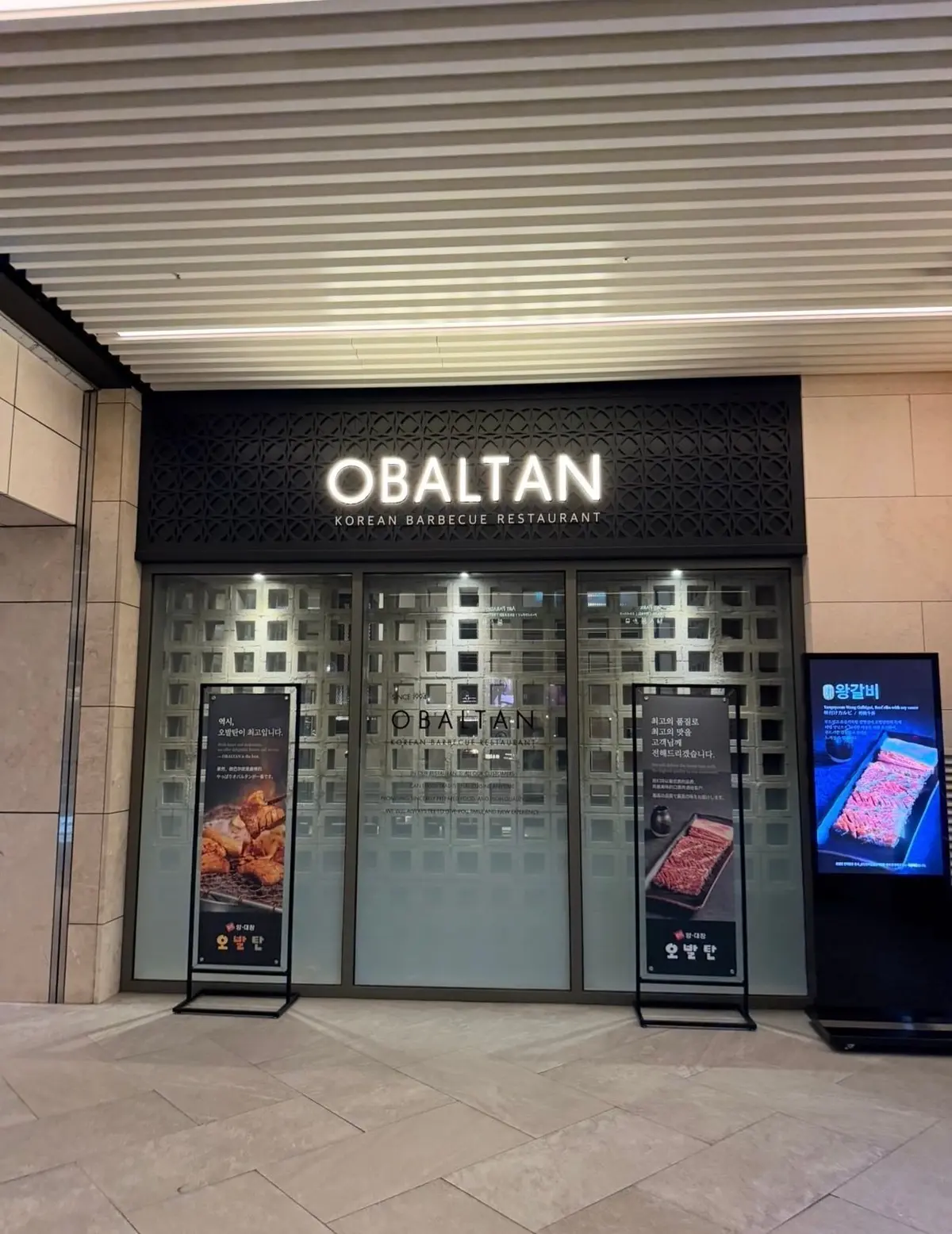 韓国料理店の「OBALTAN」の入り口、ガラスのドアと黒い装飾パネル、店名とレストラン名が書かれた看板、店外に置かれたメニューや料理写真のディスプレイが並ぶ。