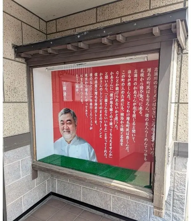 窓に貼られた縦長のポスター。中央に笑顔の男性の顔写真、下部に「三國清三」の文字。背景は赤色で、日本語の文章が書かれている。窓の前に緑色の芝生が広がっている。