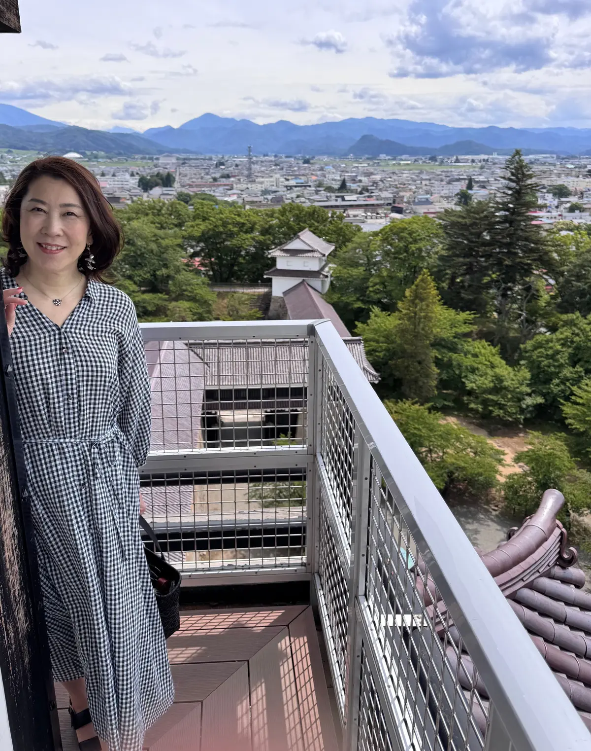 女性が屋上テラスに立ち、遠くの山並みと街並みを背景に微笑んでいる。手には黒い鞄を持ち、チェック柄のワンピースを着ている。手すりには金属製の防護柵があり、屋根の瓦が見える。