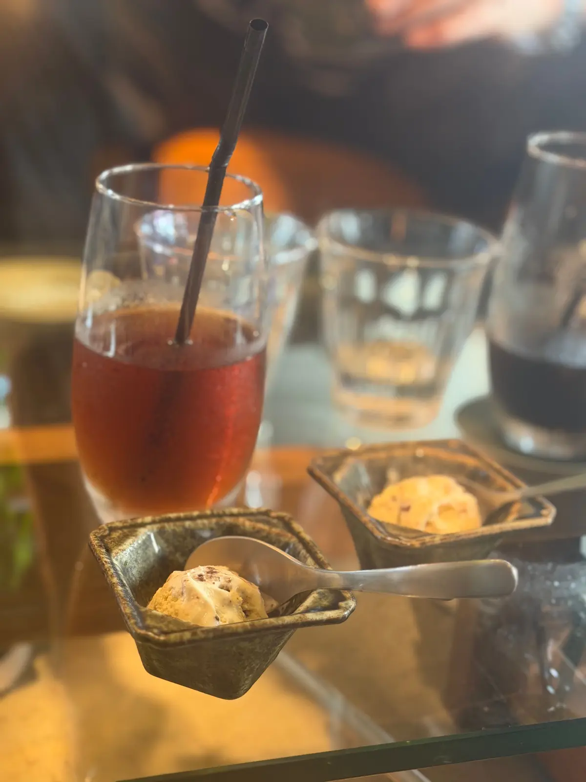 テーブルに置かれた飲み物とアイスクリーム。左には黒いストローが入った赤い飲み物、右には空のグラスとコーヒー。手前には2つの小皿があり、それぞれにアイスクリームが乗せられている。