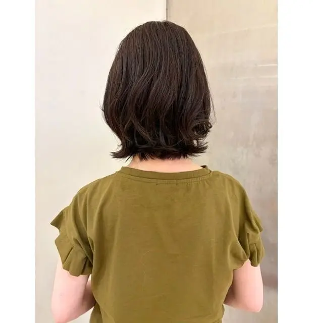 肩までの長さのダークブラウンのボブヘア。ゆるやかなウェーブがかかり、後ろ姿を捉えた写真。緑色のTシャツを着用。