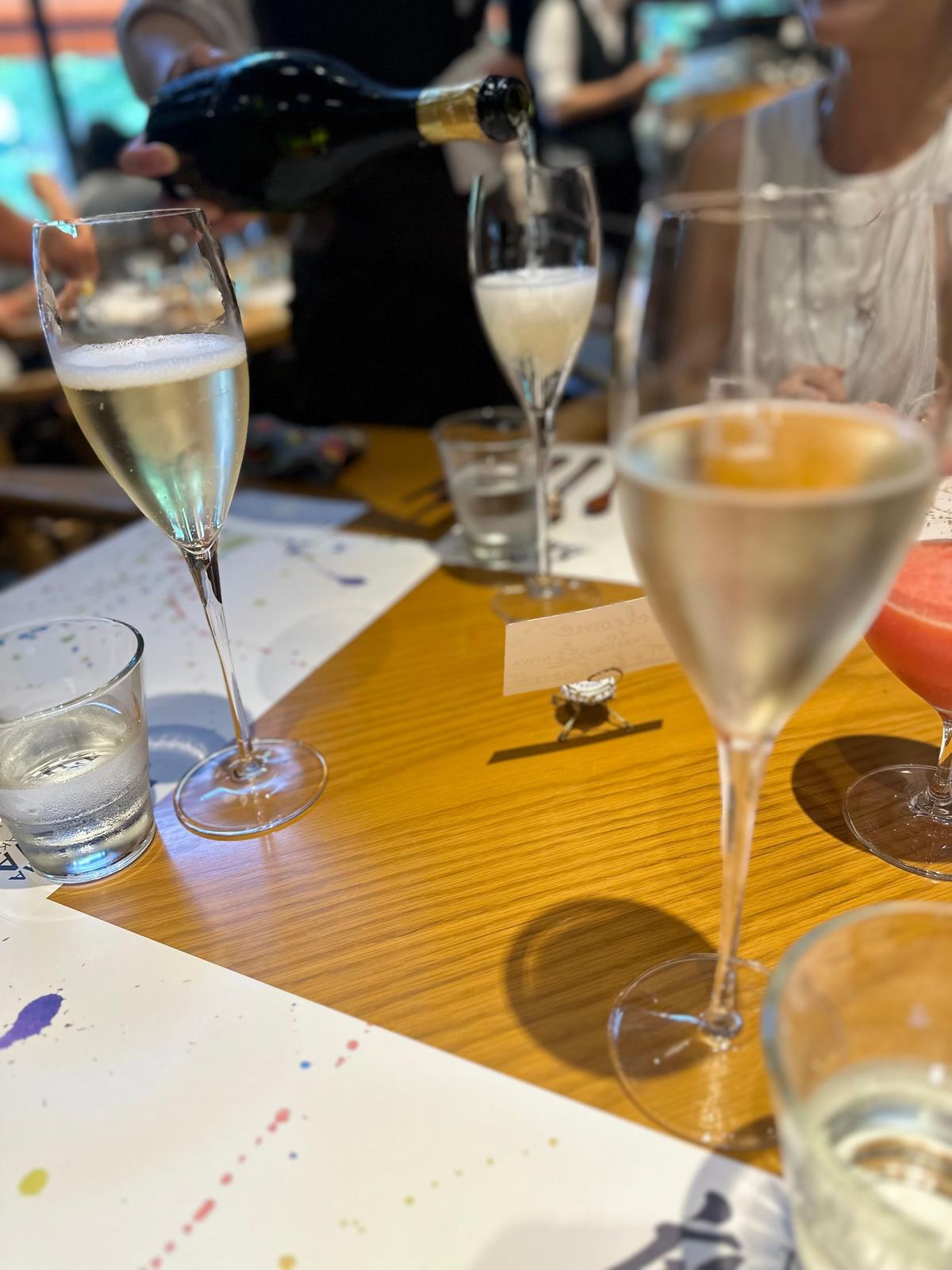 木製のテーブルに置かれた、様々な形のグラスに入った飲み物。シャンパンフルート、マルティニグラス、ロックグラスなどがあり、中には泡立つ液体や色のついた液体が見える。