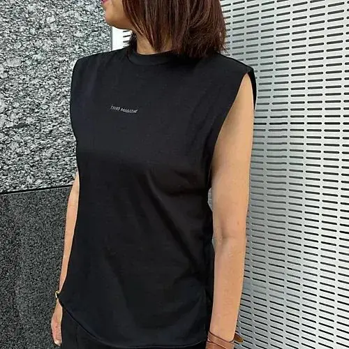 黒いカットソーを着用した女性。肩が大きく開いたデザインで、胸元に白いブランドロゴ「THIRD MAGNIFY」の刺繍が入る。茶色の腕時計とブレスレットを身に着けている。背景には石材と金属製の壁がある。