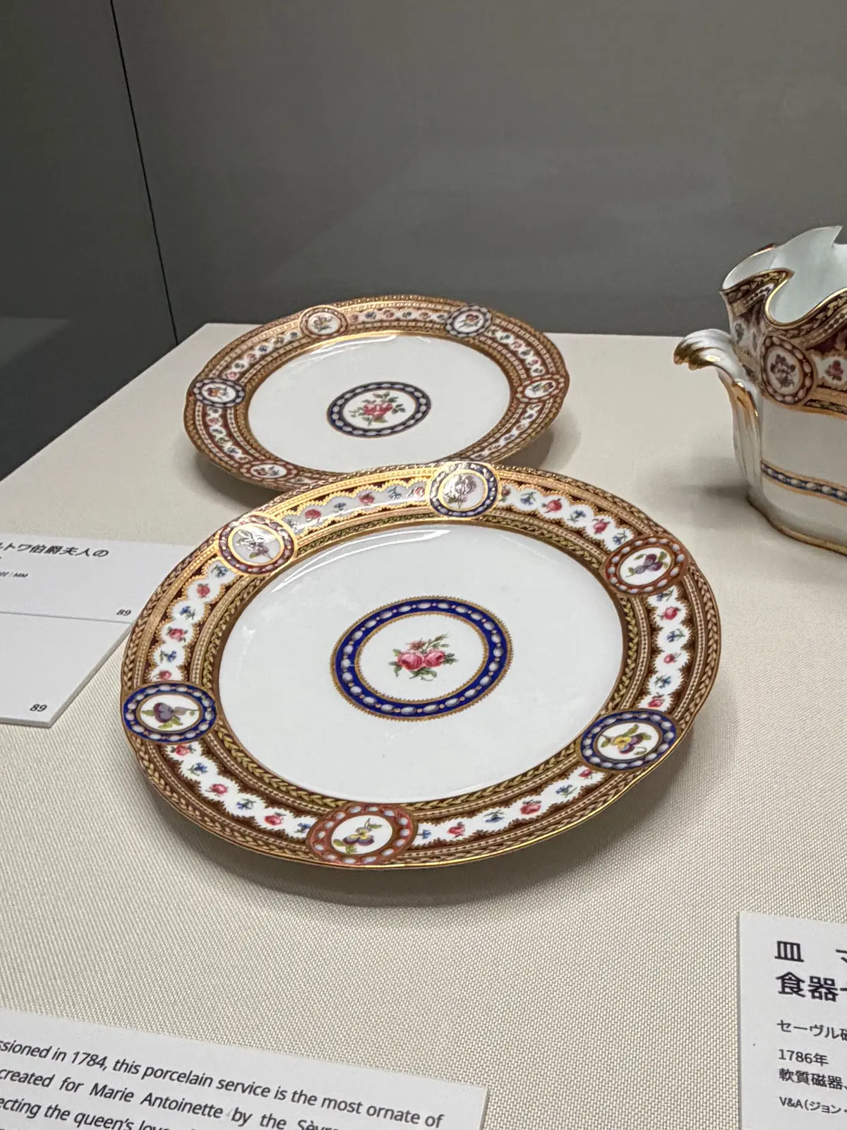 白地に金彩と赤・青・緑の装飾が施された磁器の皿2枚と、同じ模様のミルクピッチャーが置かれている。皿は積み重ねられ、背景には説明書きが見える。