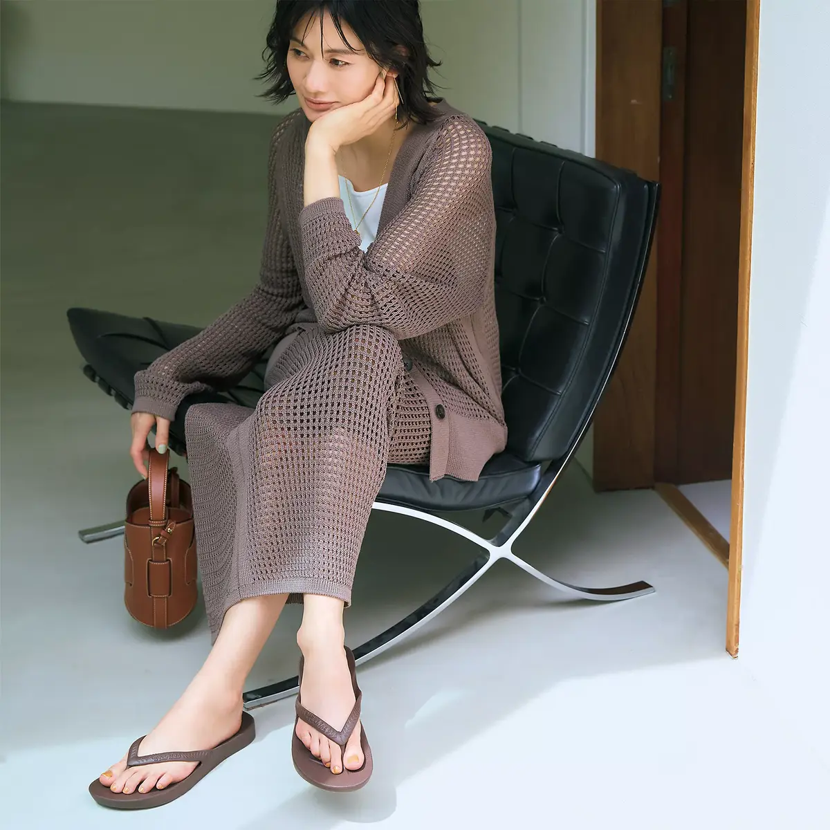 茶色のニットカーディガンとロングスカートを着用した女性が、黒い革張りの椅子に座っている。茶色のサンダルを履き、手に茶色のバッグを持っている。背景は淡い緑色の壁。