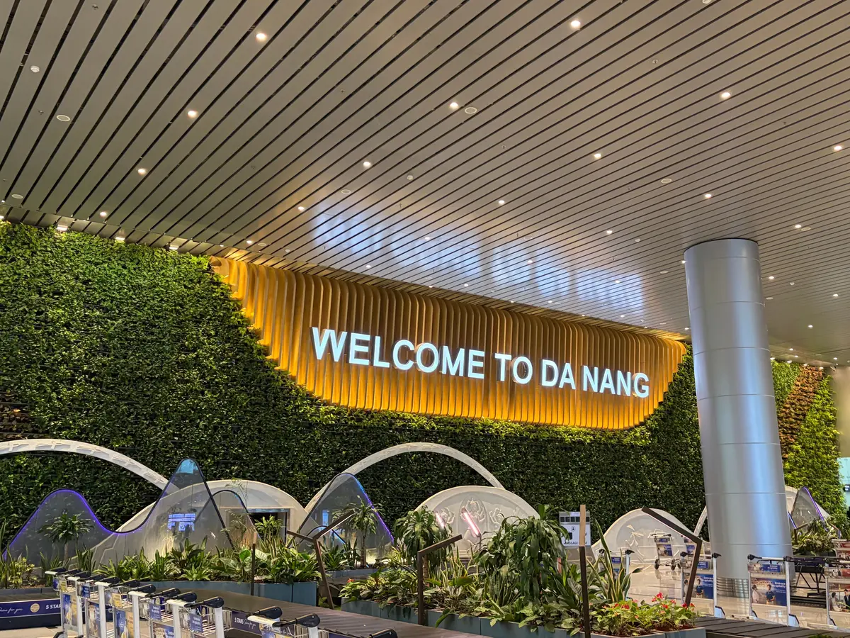 空港の到着ロビーに設置された、「WELCOME TO DA NANG」のネオンサイン。背景には緑の壁面緑化が広がり、手前にはトロリーや観葉植物が配置されている。天井は水平方向にラインが並んだ構造。