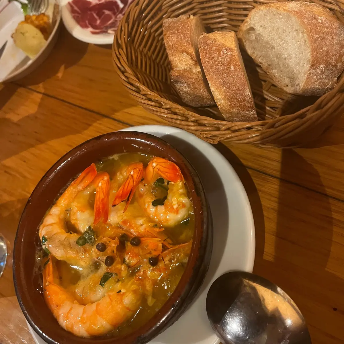 木製のテーブルに置かれた、小鍋に入ったエビの料理。エビはオレンジ色で、カパーやハーブが添えられている。右にはパンの籠、奥には前菜の皿が見える。
