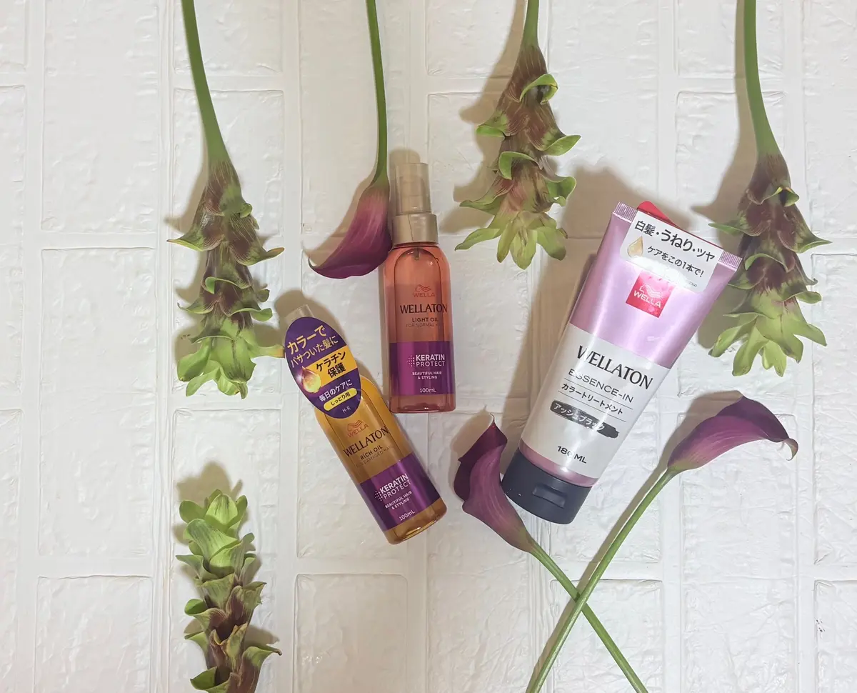 ウェラ(Wella)のヘアケア製品（オイル、エッセンス、マスク）が、紫色の花と緑の葉に囲まれて置かれている。白い背景。