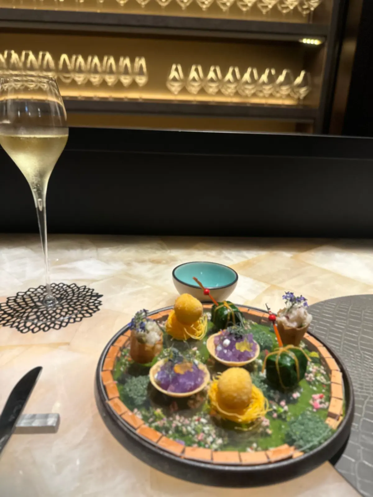 丸いプレートに、緑の葉や花で飾られた小さな料理が並ぶ。中央に黄色いボールが置かれ、周囲に紫の装飾、緑の野菜、小さな花が添えられている。プレートの横にはワイングラスとナイフが置かれている。