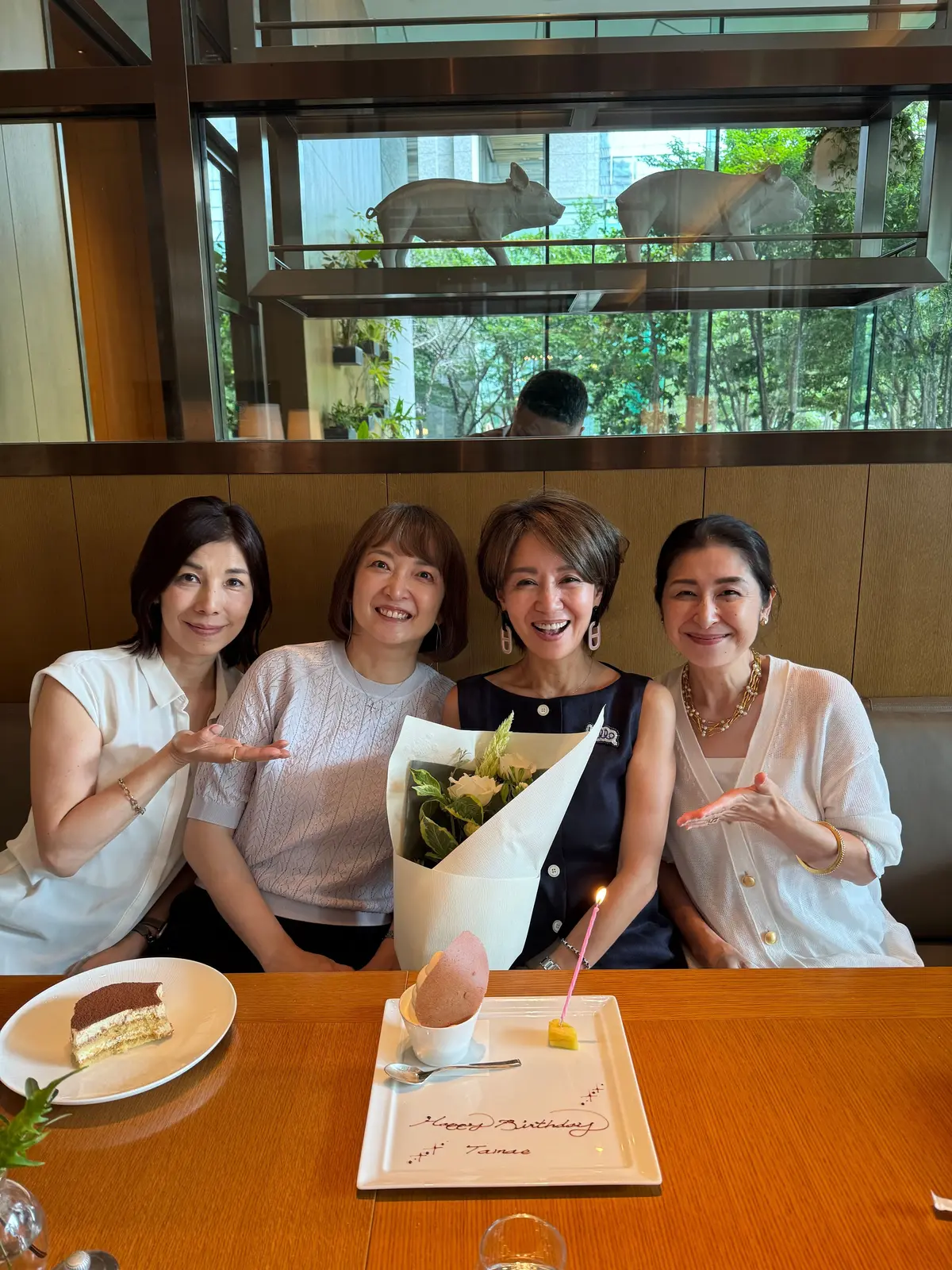 レストランで誕生日のお祝いをする4人の女性。中央にろうそくのついたケーキがあり、手前にチョコレートケーキの皿。背景には猫の置物が飾られた大きな窓。