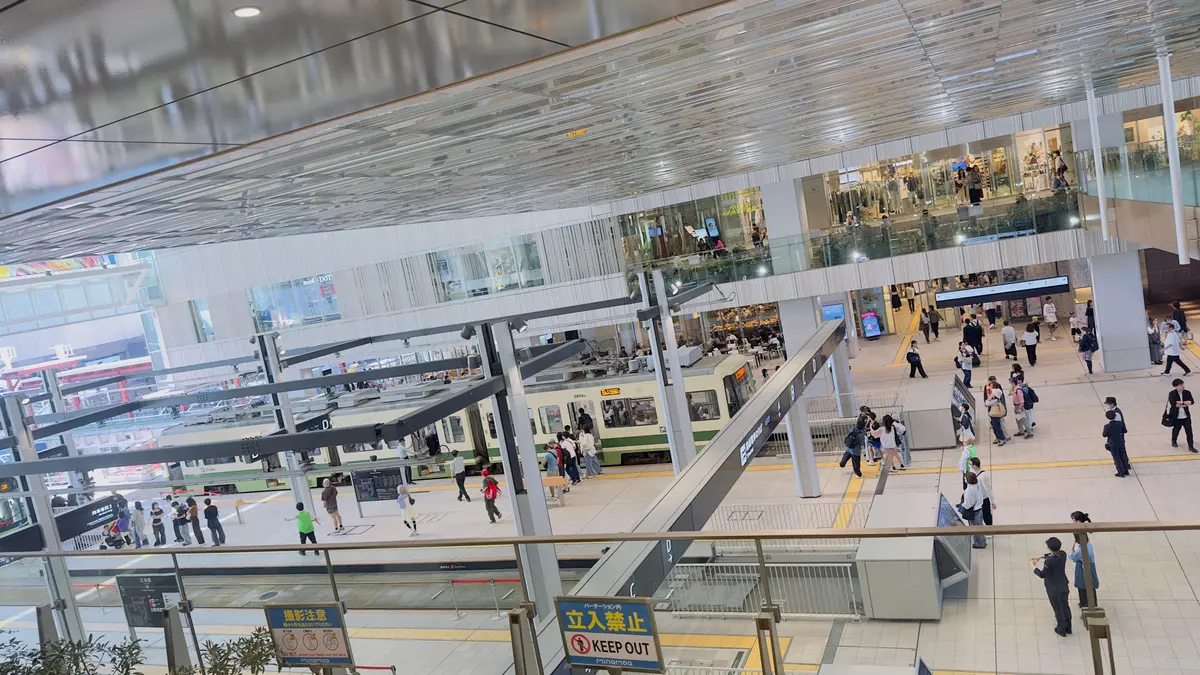 駅の中央に電車が停まり、多くの乗客がプラットフォームを歩いています。ガラス張りの天井とエスカレーターがあり、周囲にはショッピングエリアが広がっています。