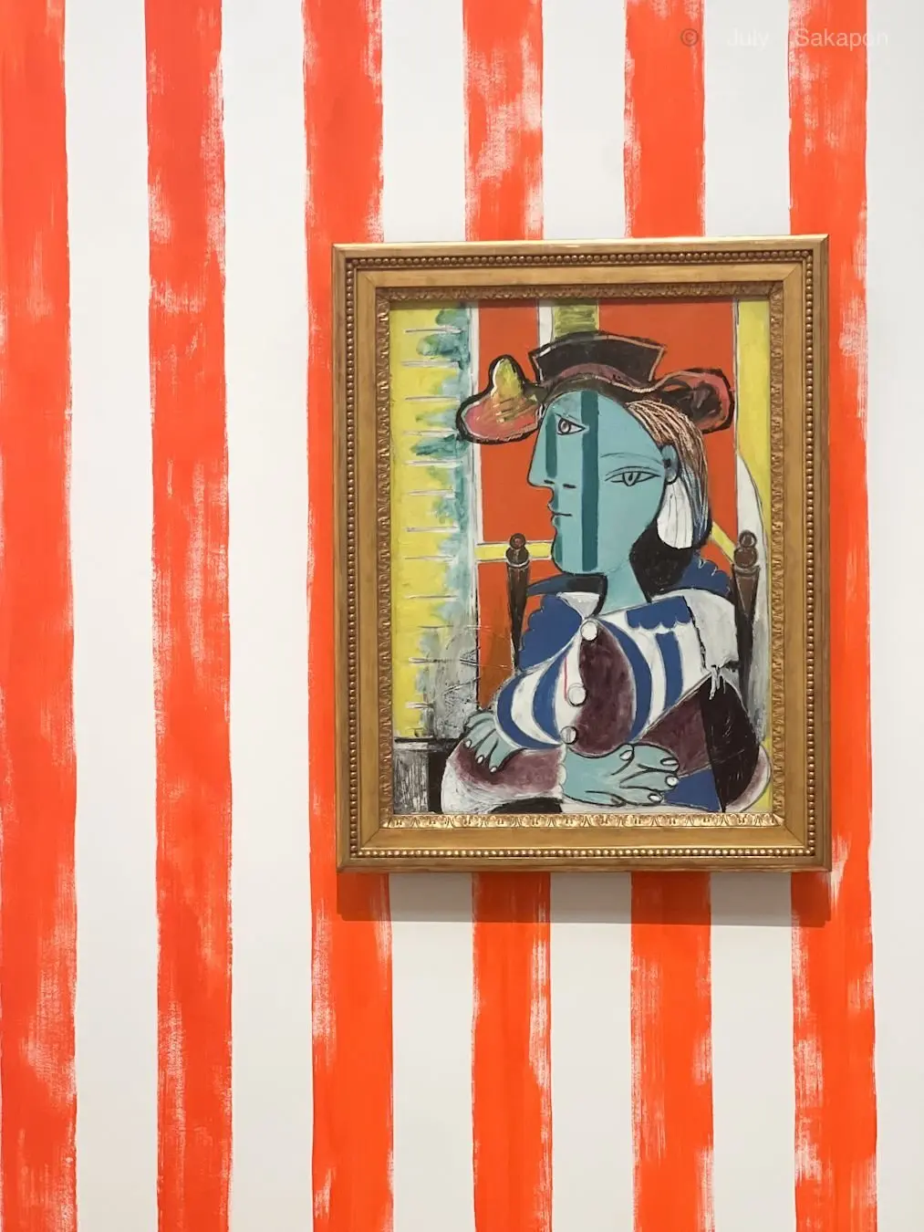 青い肌の男性が金色の額に入った絵画、赤と白のストライプ背景に飾られている