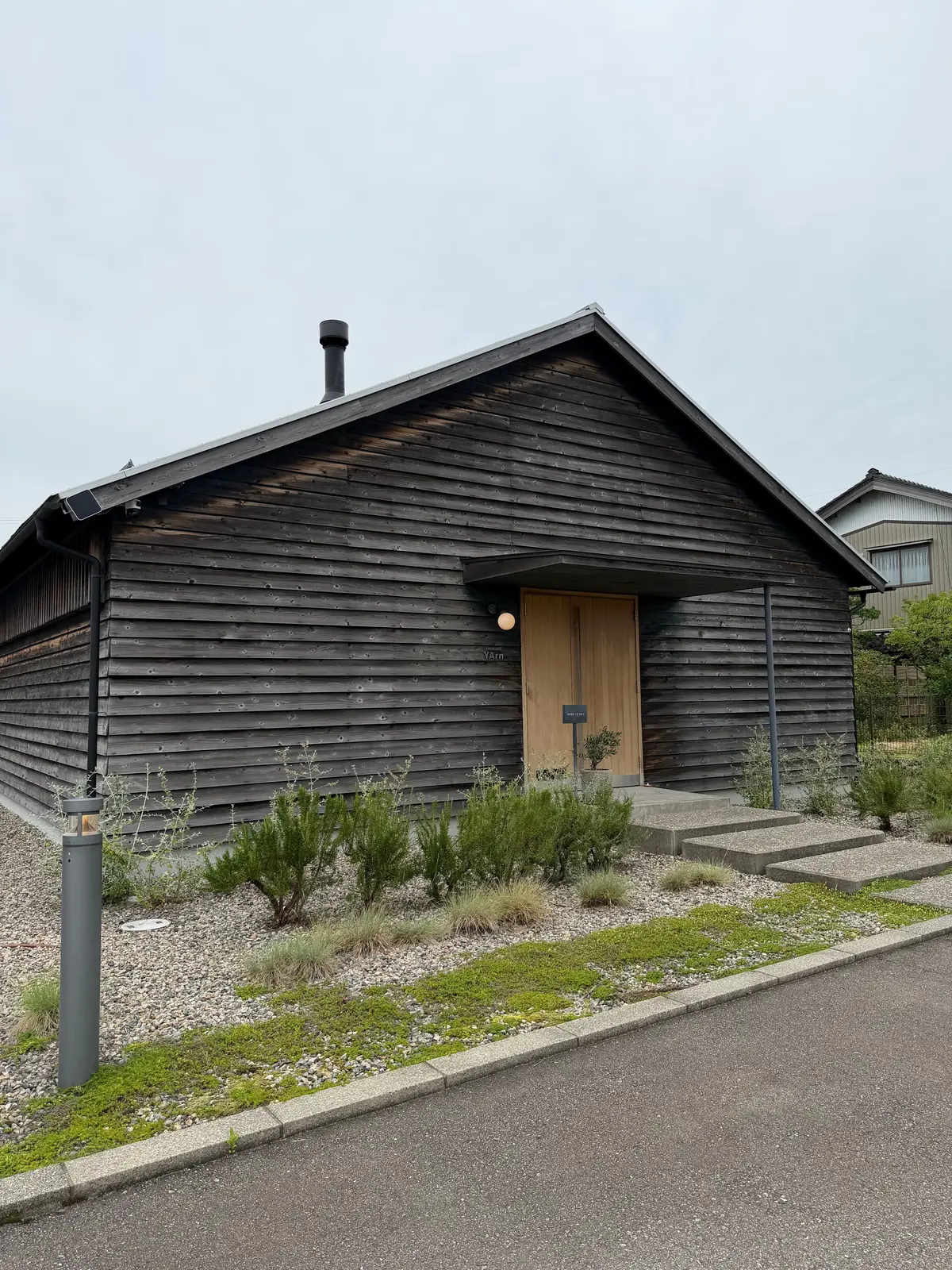 木製の板張りの外観を持つ、シンプルな小屋。正面には木製の扉があり、左右に照明が設置されている。小屋の周囲には雑草が生えた小石敷きの庭。背景には別の家が見える。