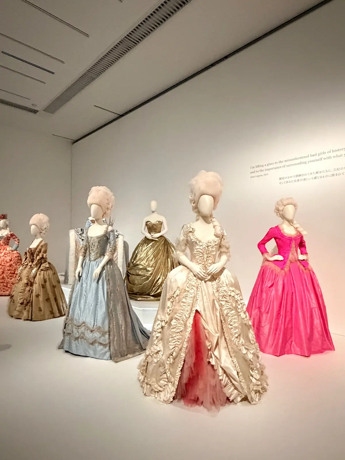 展示ケースに並んだ、18世紀風の豪華なドレスを身につけたマネキン人形。色とりどりのドレスが並び、ピンク、ゴールド、水色などが目立つ。背景には白い壁と天井。