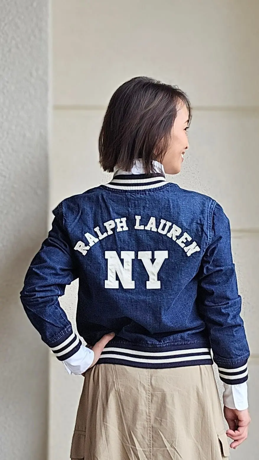 紺色のデニム素材のラルフローレンのボンバージャケットを着用した女性。背面に白字で「RALPH LAUREN」と「NY」の文字が刺繍されている。ベージュ色のスカートを着用し、白い襟が見える。