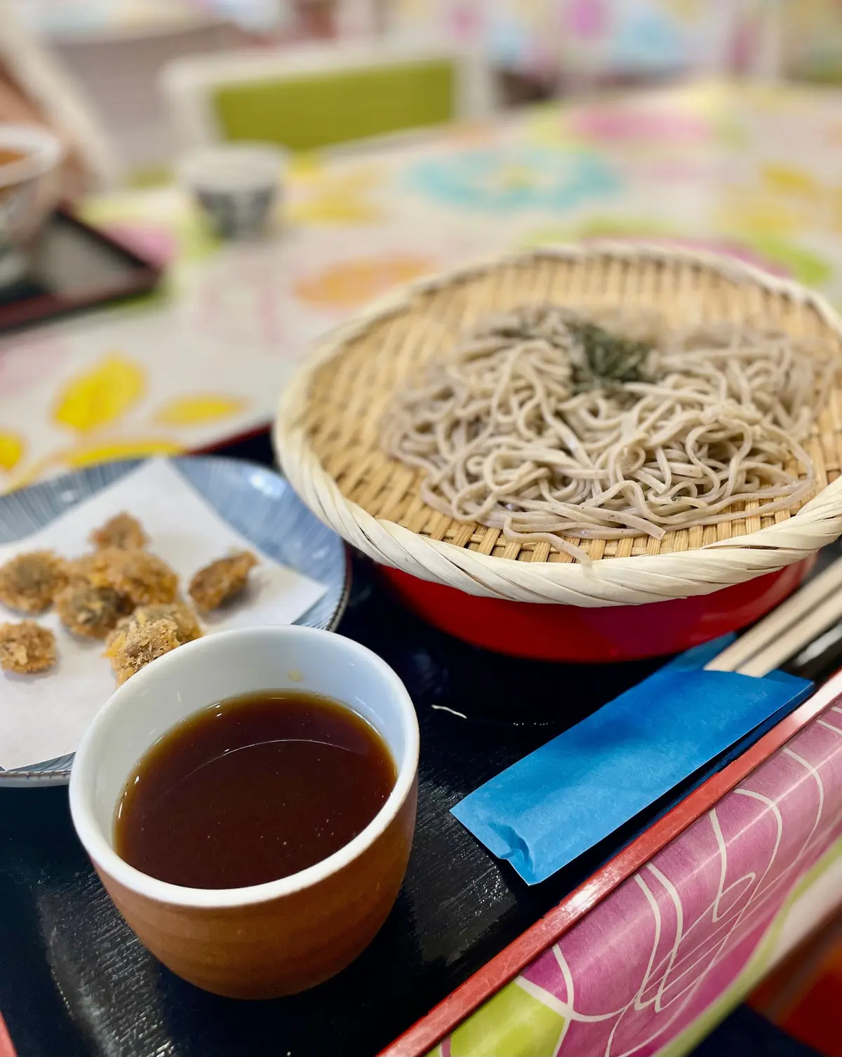 竹製の器に盛りられた細麺と、茶色のソースが入った白い小皿、揚げ物の盛り合わせが並ぶ料理のセット