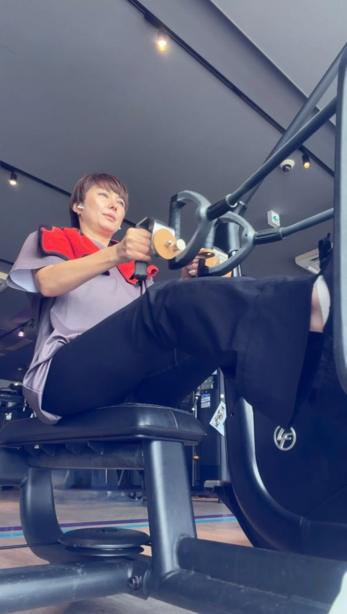フィットネスジムで、黒いパンツと白のシャツを着た女性が、EMS（電気筋肉刺激）トレーニングマシンを使用している。女性は真剣な表情で、マシンに掴まり、片足を上げて運動している。背景には、他のトレーニング機器が見える。