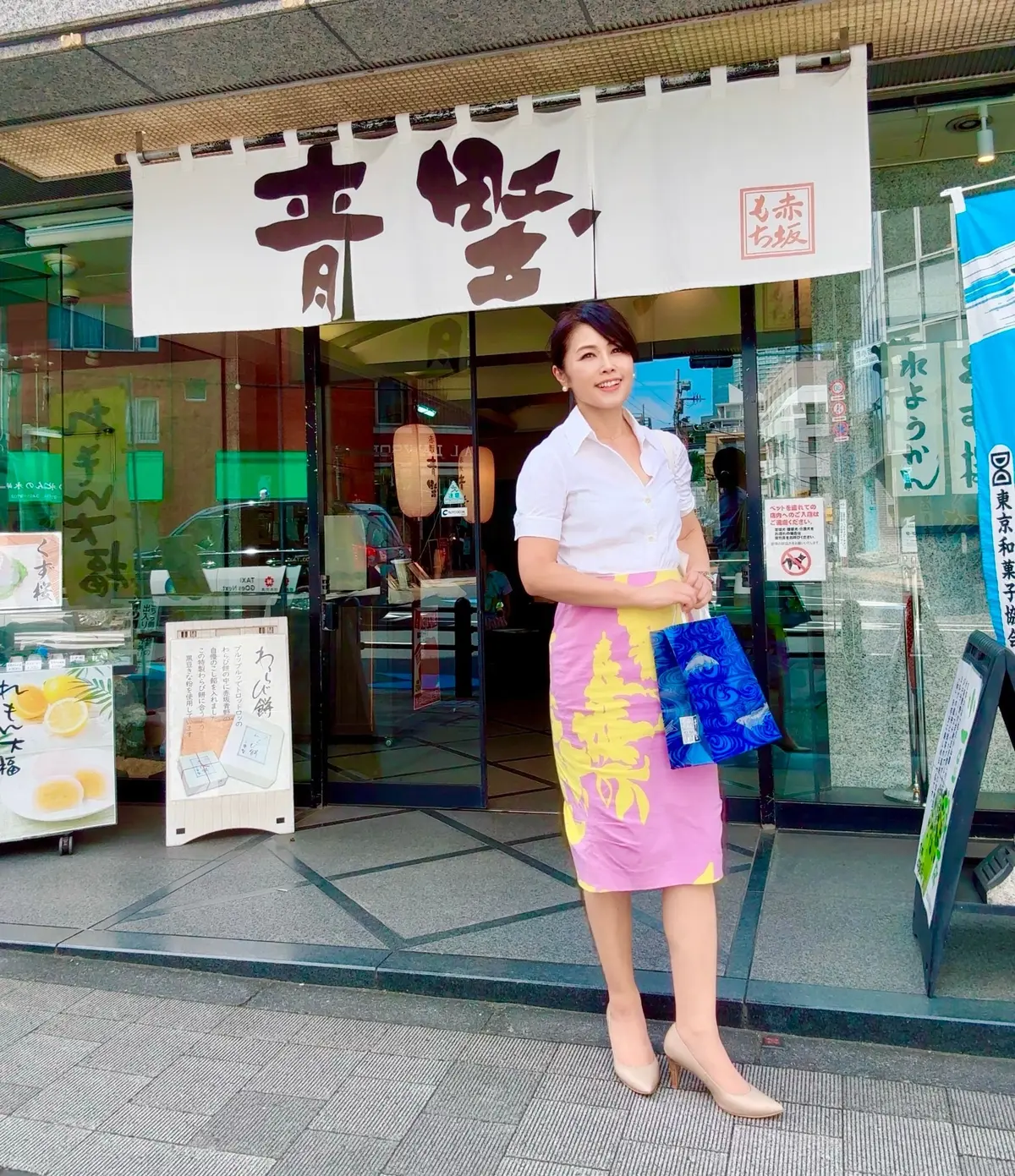 白いシャツに花柄のミニスカートを着用した女性が、日本風の店の前で微笑んでいる。右手に青い扇子を持っている。店の看板は黒地に白文字で「青柳」と書かれている。