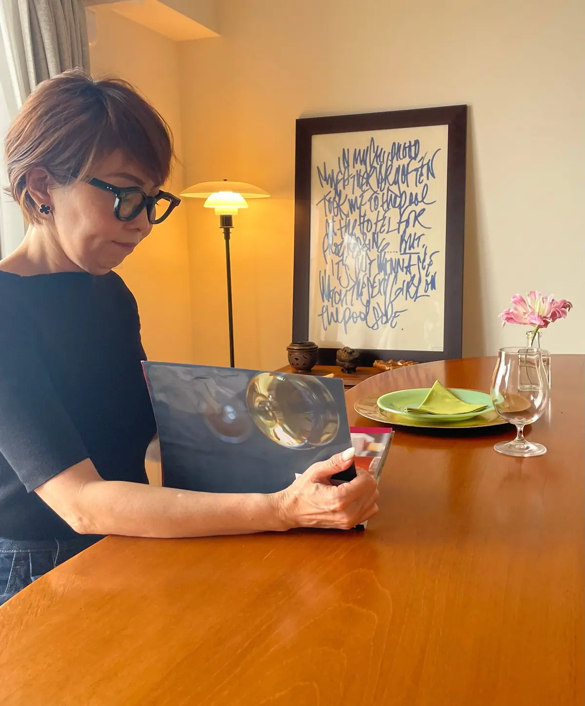 メガネをかけた女性が、丸い模様が描かれた青い額装された絵画を手に持つ。背景には、花の入った花瓶、ワイングラス、緑のプレートが置かれた木製のテーブルと、壁に飾られた額装された文字が並ぶ。