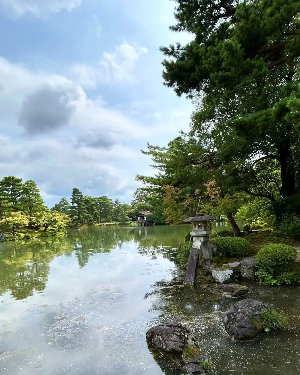 穏やかな水面が広がり、緑豊かな木々と石組みが配置された日本庭園。中央には灯籠が置かれ、背景には建物が見える。雲が浮かぶ青空。
