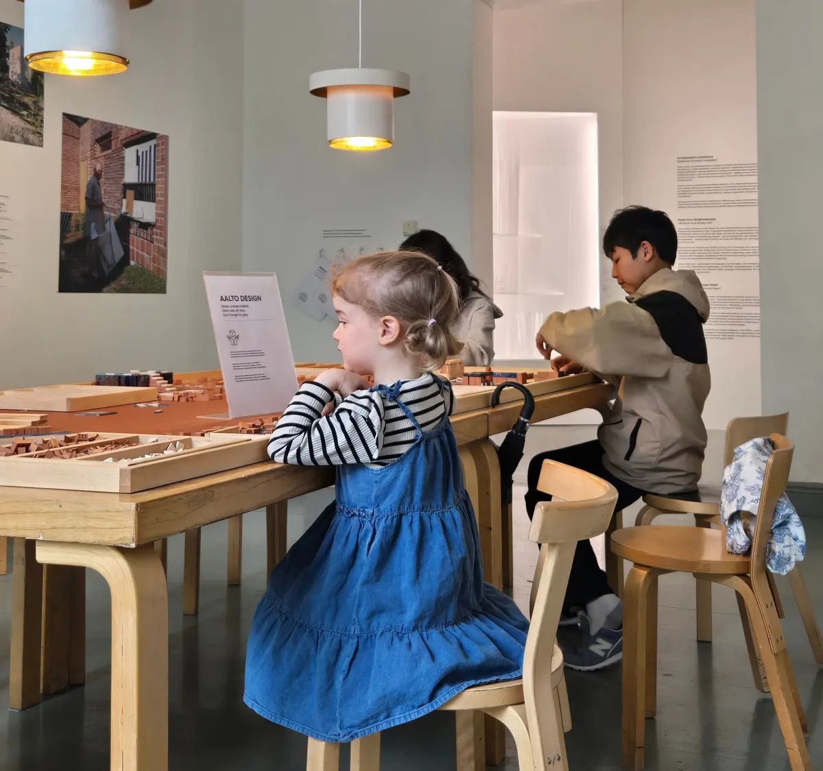 白壁の展示室で、木製のテーブルを囲む子供たち。左には円形のペンダントライト、右には白いペンダントライトが吊り下げられている。