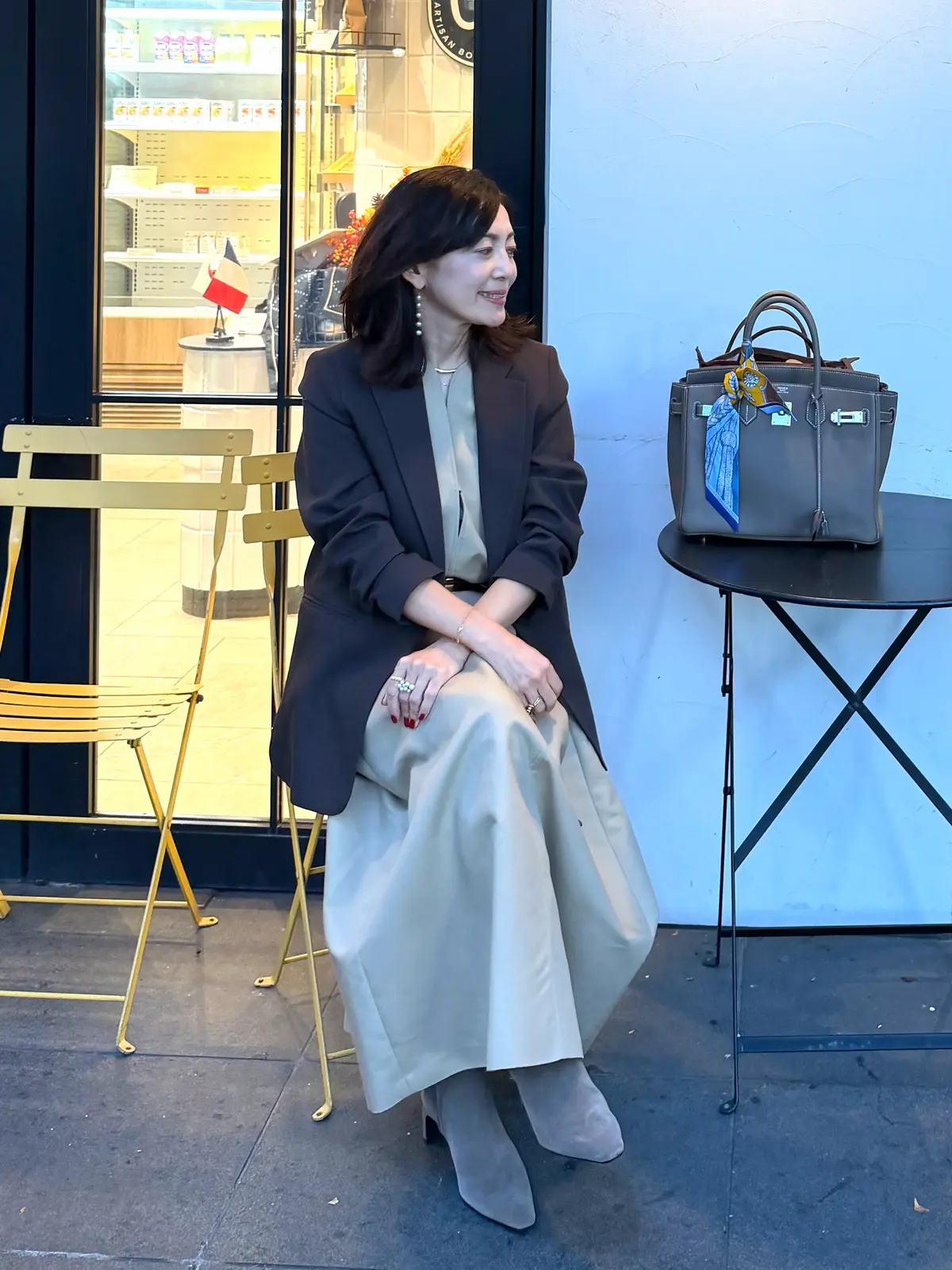 黒いジャケットとベージュのフレアスカートを着用した女性が、屋外のベンチに座っている。右手にハンドバッグを持ち、視線を下げている。背景には店舗の窓と、テーブルと椅子が見える。