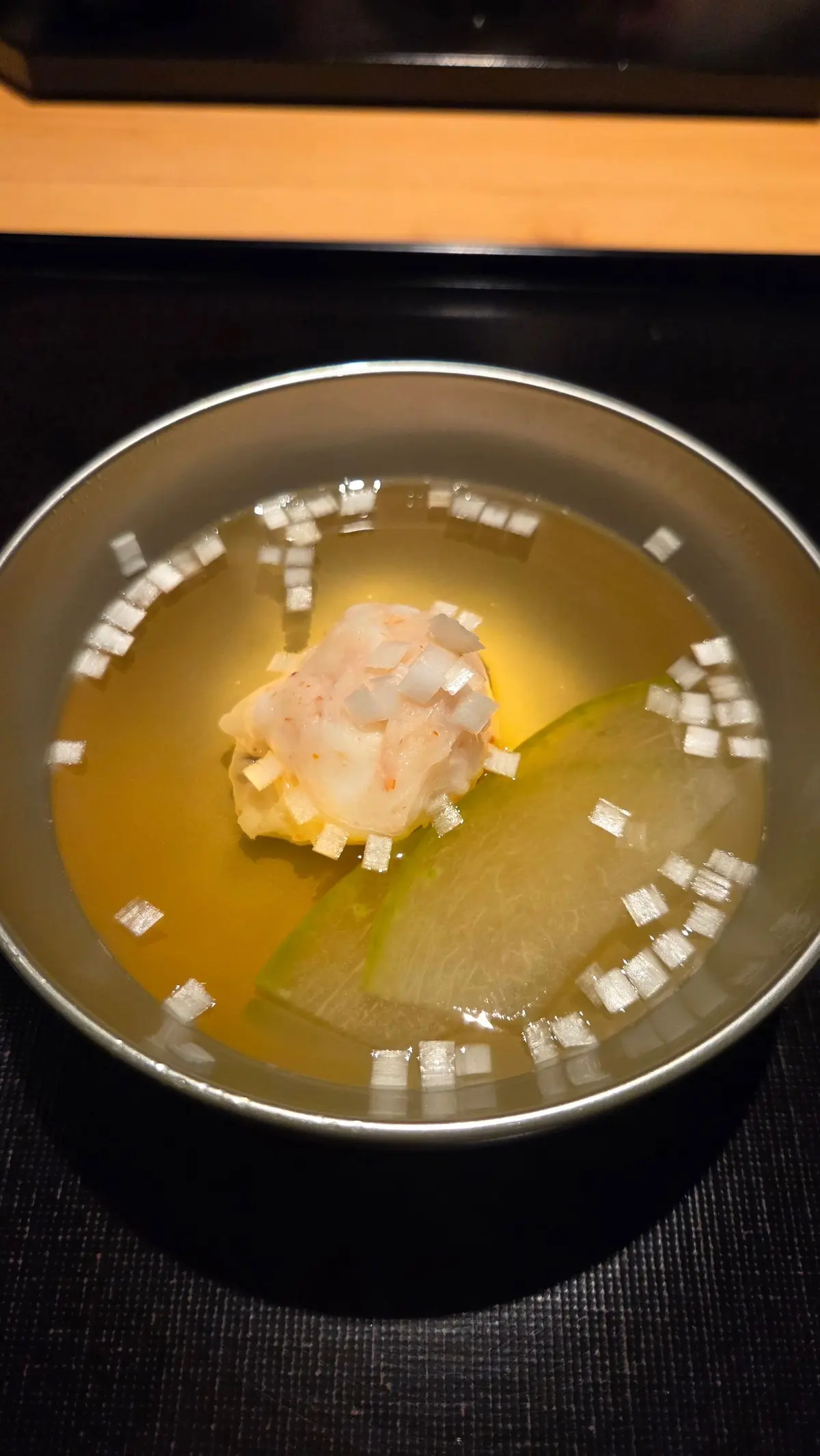 灰色の器に入った、薄い黄色いスープ。中央に淡いピンク色の魚の切り身、緑色の薄切り野菜、白い角切りが散らばっている。