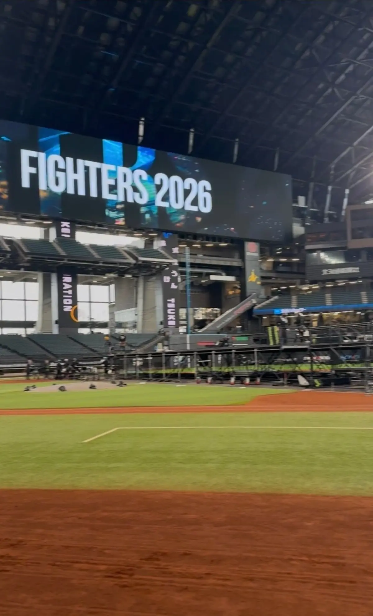 屋根付きの野球場で、フィールドには緑の芝生が広がっている。背景には「FIGHTERS 2026」の文字が表示された大型スクリーン。観客席には座席が並び、一部に金色の装飾がある。