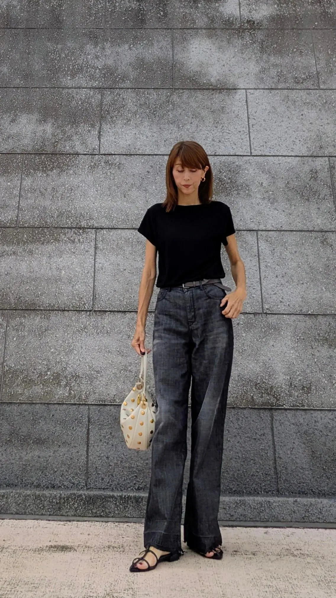 女性がグレーの石造りの壁の前で立っている。黒の半袖Tシャツ、グレーのワイドパンツ、黒のサンダルを着用。右手に白のハンドバッグを持っている。