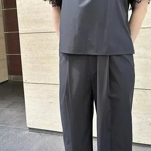 グレーの無地トップスとワイドパンツを身に着けた女性が、ベージュの壁に寄りかかった姿勢で立っている