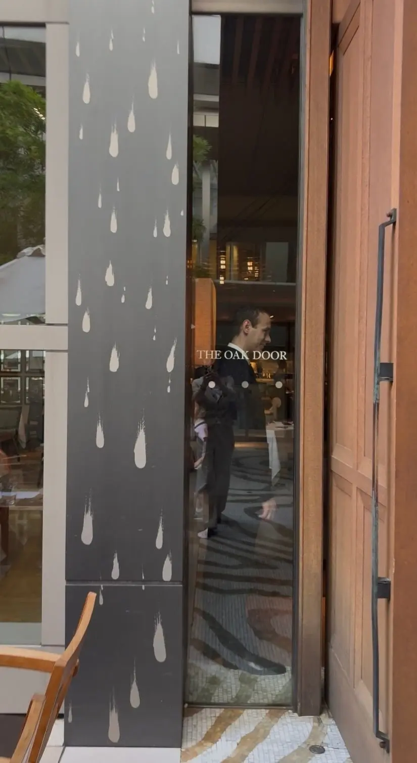 灰色の柱に白い雨粒の模様が描かれた外観。ガラス戸には「THE OAK DOOR」のロゴと、入店する男性の姿が映っている。