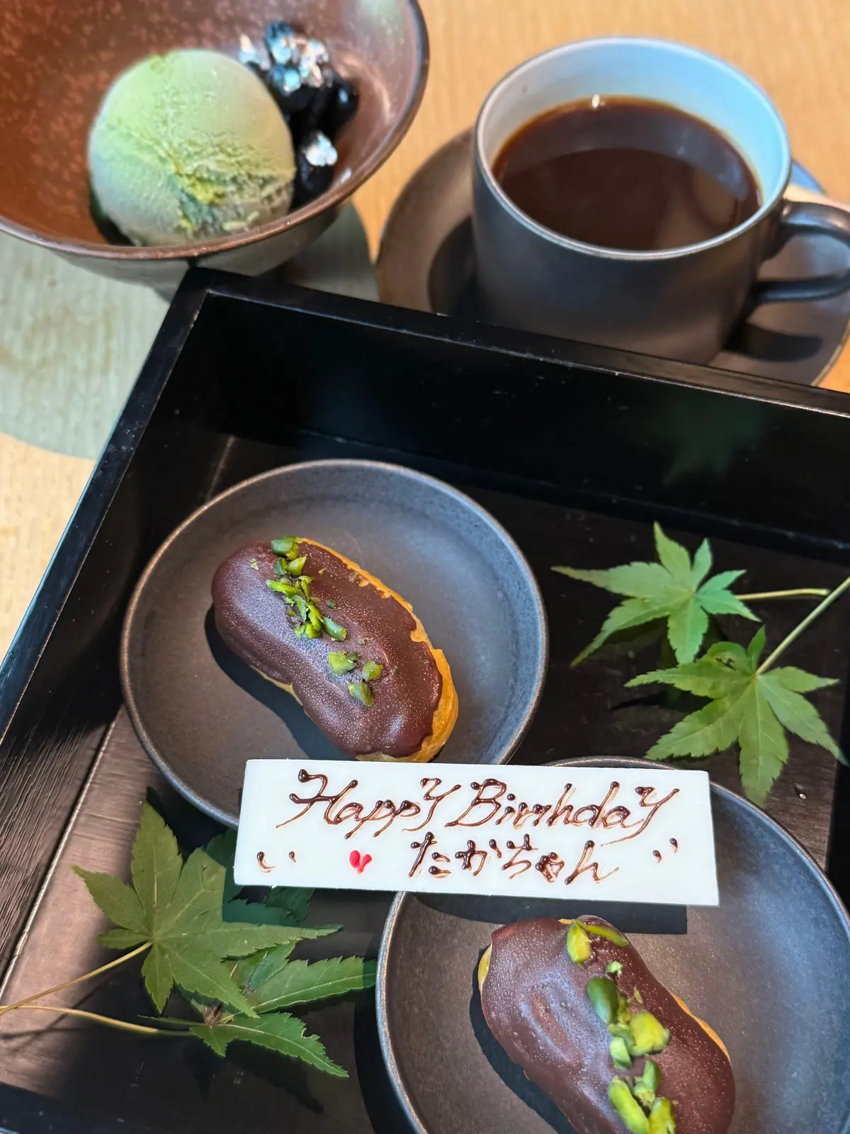 黒い木製のトレイに、抹茶アイス、コーヒー、赤茶色の丸いお菓子が盛り付けられている。お菓子には緑色のものが散りばめられ、葉っぱが添えられている。白いカードには「Happy Birthday」と名前が手書きで書かれている。