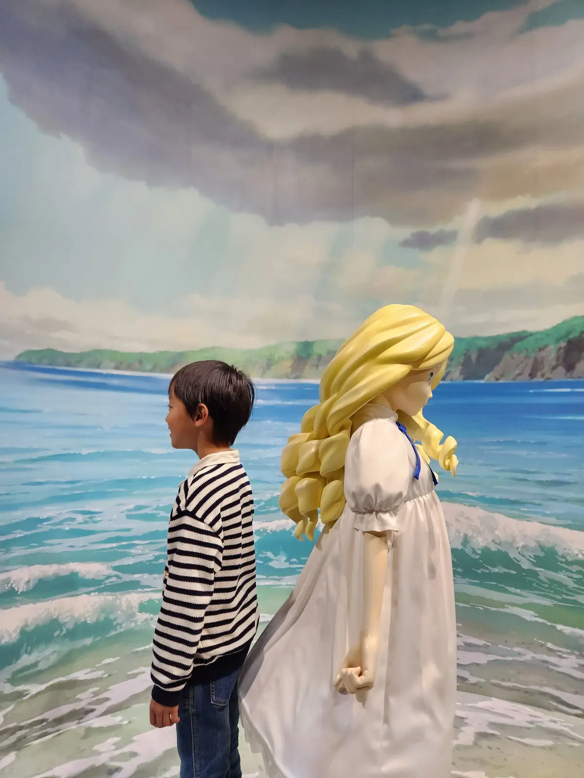 海を背景に、ボーダーのシャツを着た男の子と、金髪の大きなマネキン人形が並んで立っている。マネキンは白いワンピースを着ている。