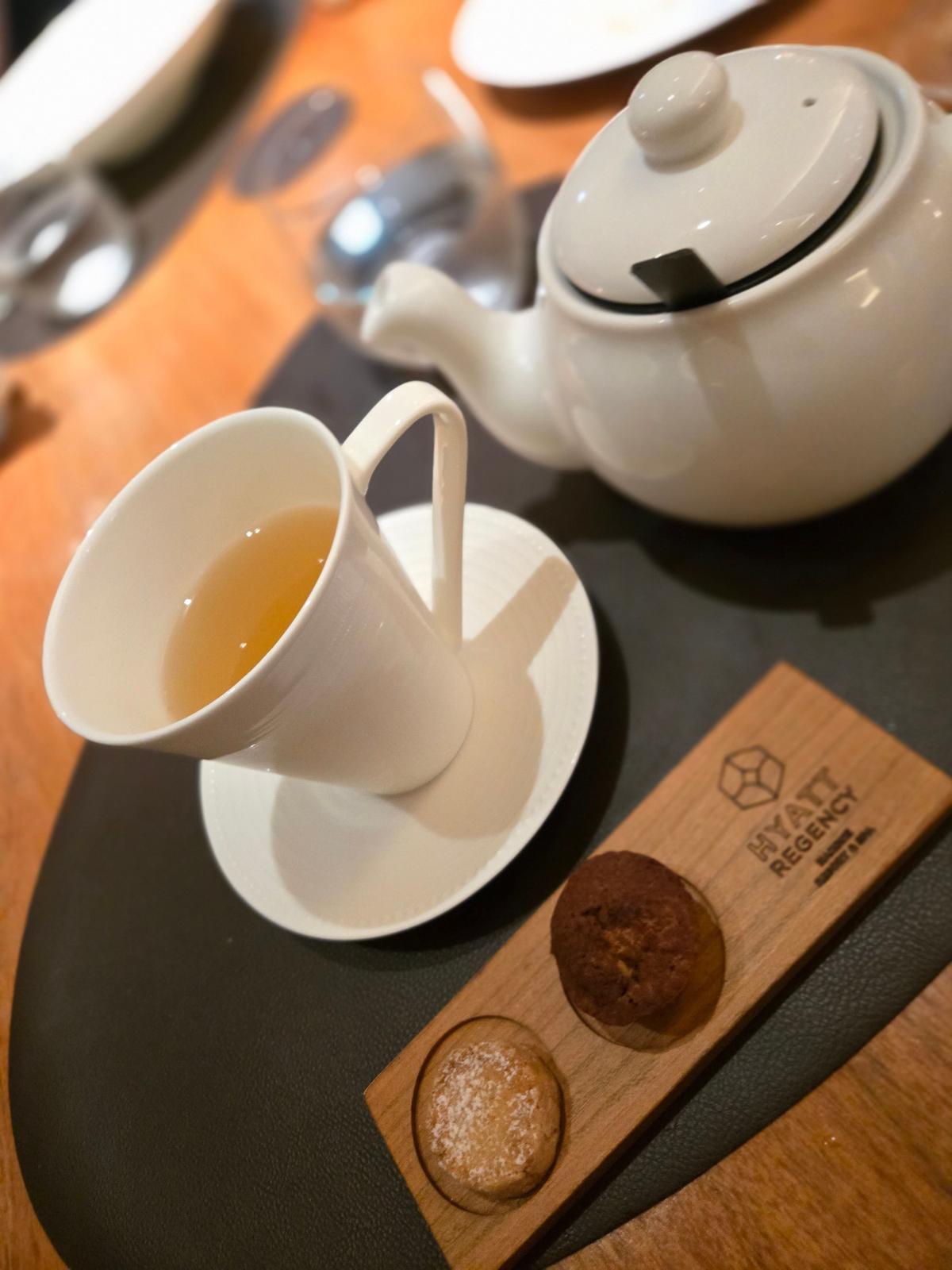 白いお皿に注がれた緑茶と、そばに置かれた急須。木製のプレートには、2つの焼き菓子と「Hyatt Regency」のロゴが刻まれている。