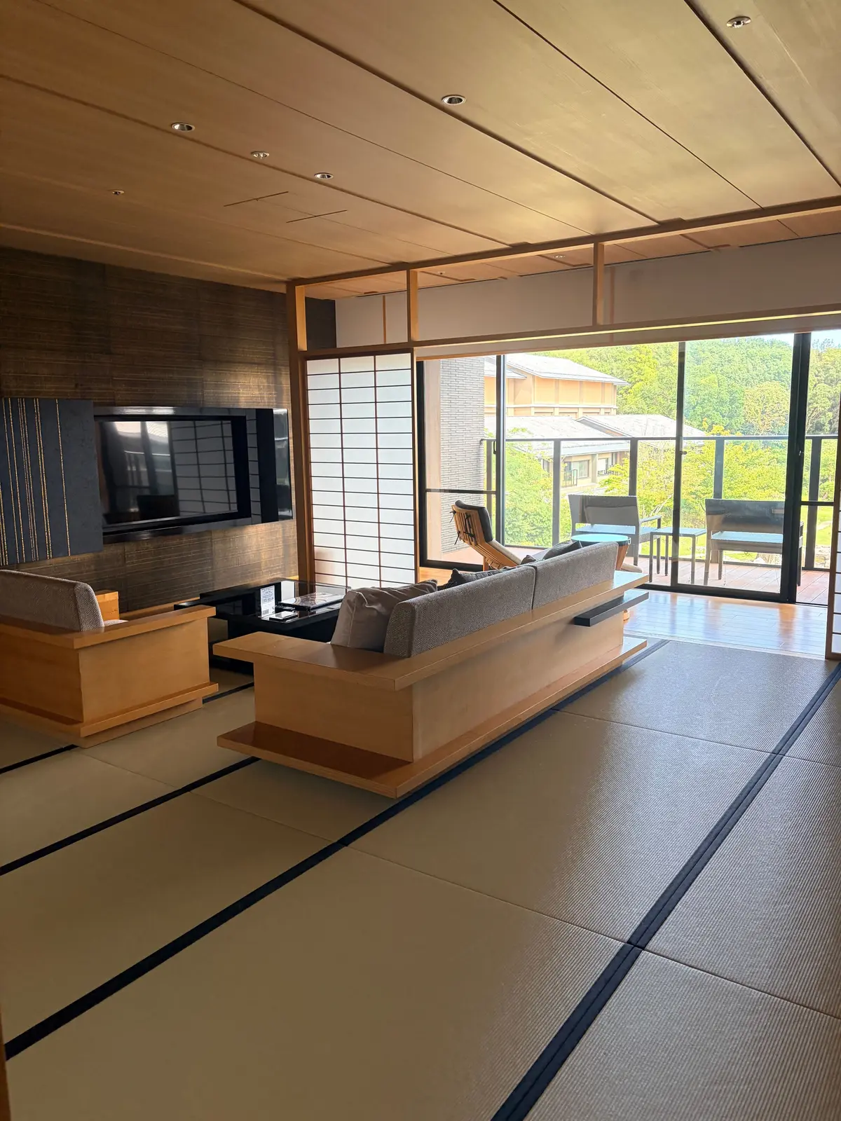 木目調の天井と畳敷きの広々としたリビング。木製のソファとローテーブル、壁掛けテレビが配置。大きな窓から緑豊かな景色が見える。