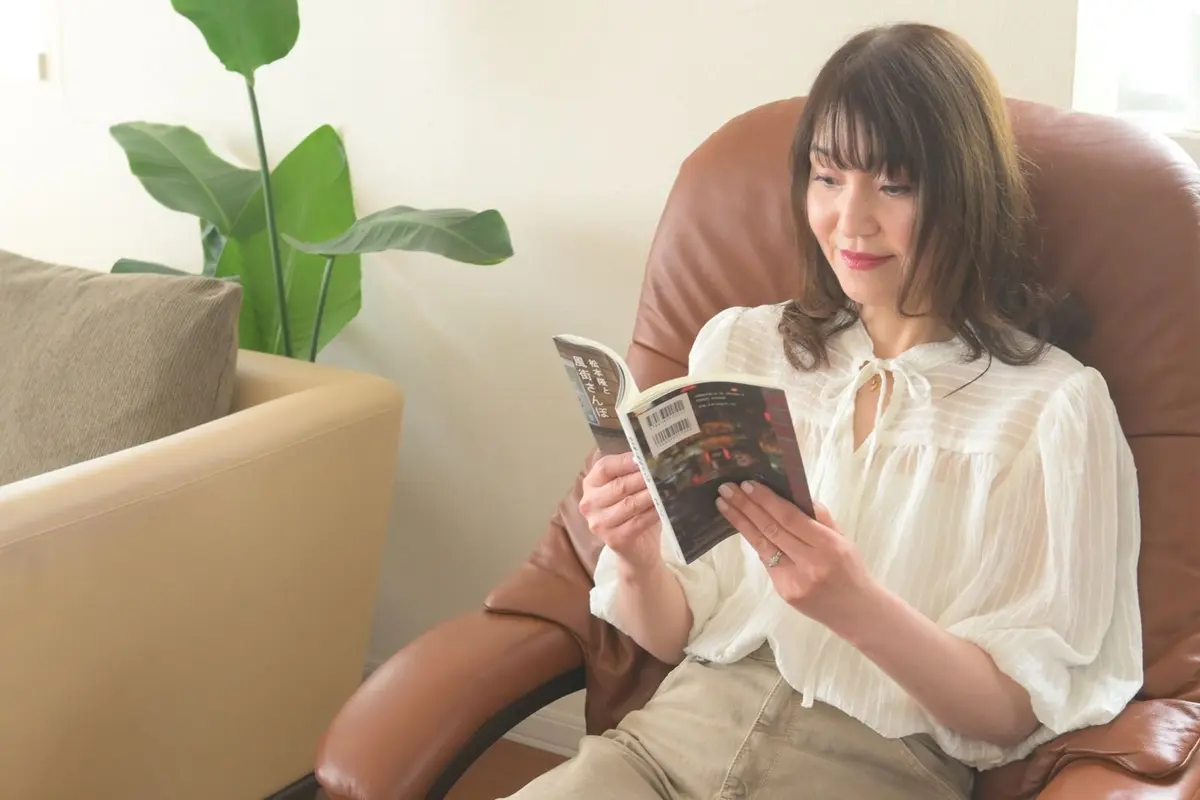 女性が茶色のレザーソファに座り、本を読み、背景に緑の観葉植物が置かれている