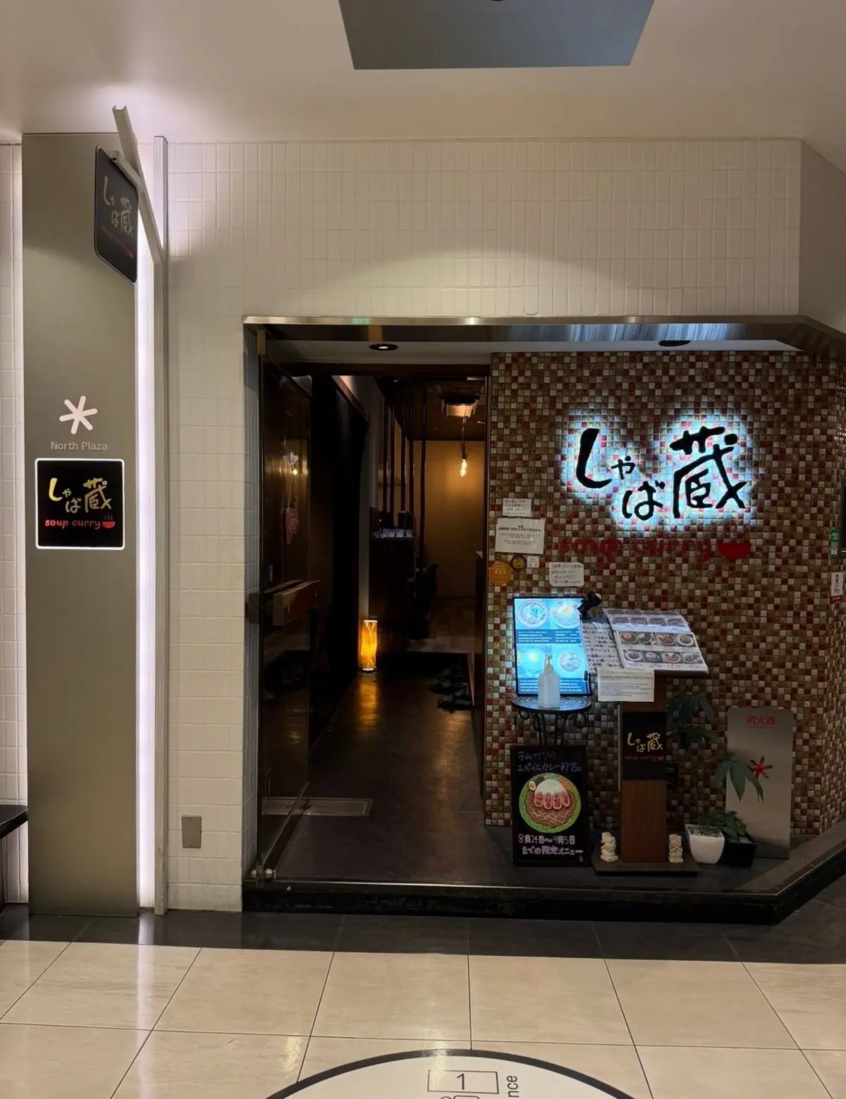ショッピングモールの北側の入り口に面した、カレー専門店「じゃばば」の店舗外観。木目調の看板と、メニューや装飾品が置かれた入り口付近。店内は薄暗く、テーブル席が見える。