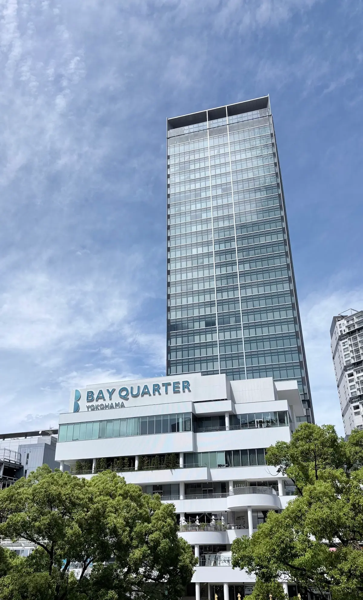 **BAY QUARTER YOKOHAMAと記載されたガラス張りの高層ビルが、青空の下に緑の木々を背景に立つ**