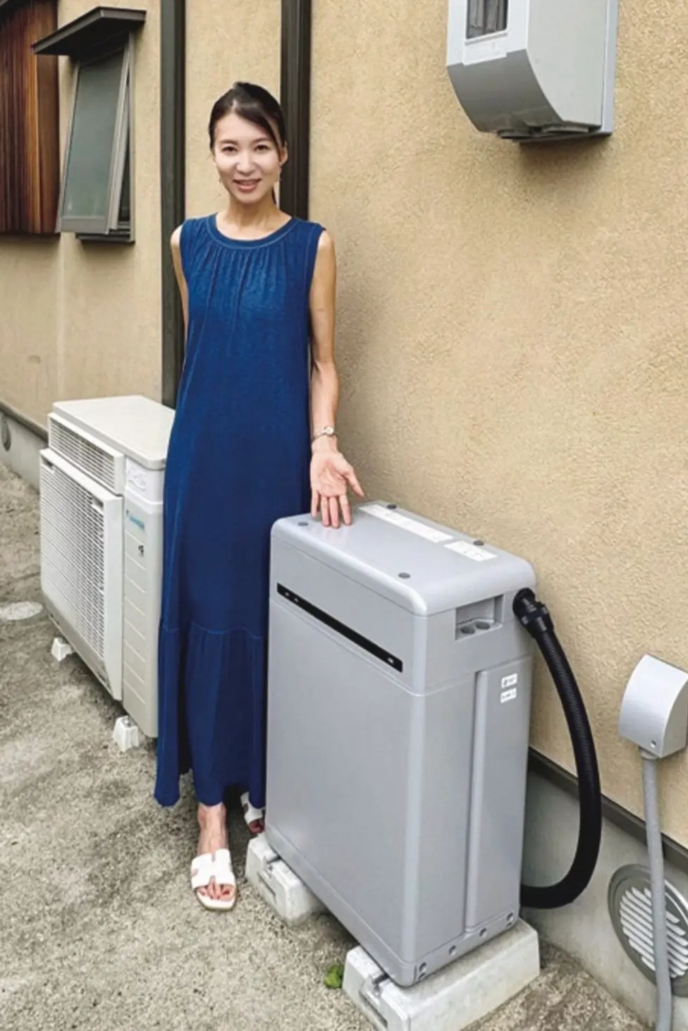 青いワンピースを着た女性が、グレーの壁に設置された大型の機械の横に立っている。機械の周りには黒いホースや白い排気口が見える。背景には窓とエアコンの室外機がある。