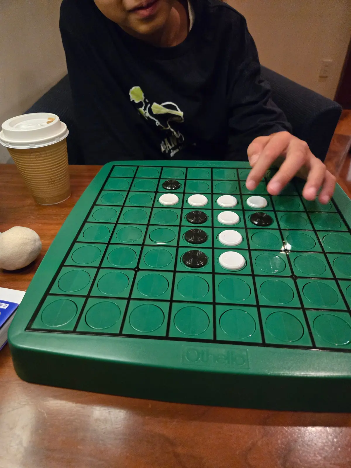 Othello（オセロ）のゲーム盤に、黒と白の石が配置されている様子。少年が石を置こうとしており、背景には飲み物のカップと白い石が見える。