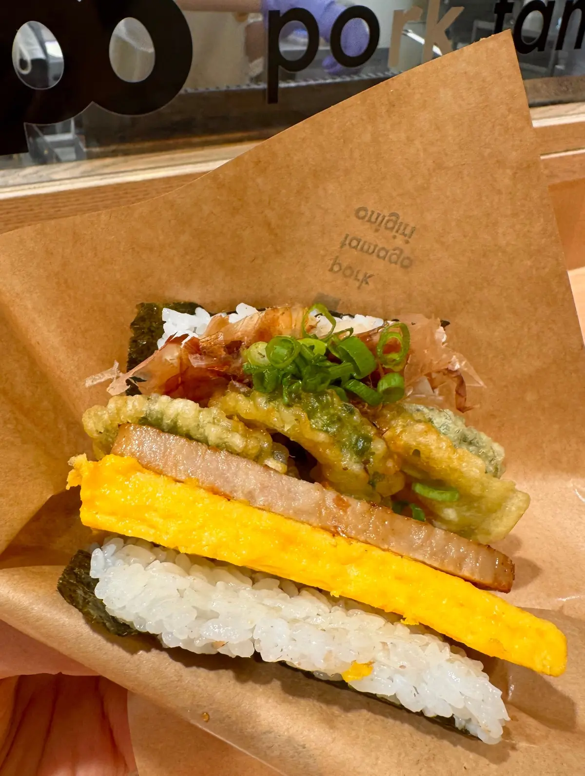 紙の包み紙に盛り付けられた、米と野菜、肉、揚げ物をトッピングした料理。黄色の野菜と緑のネギが目立つ。