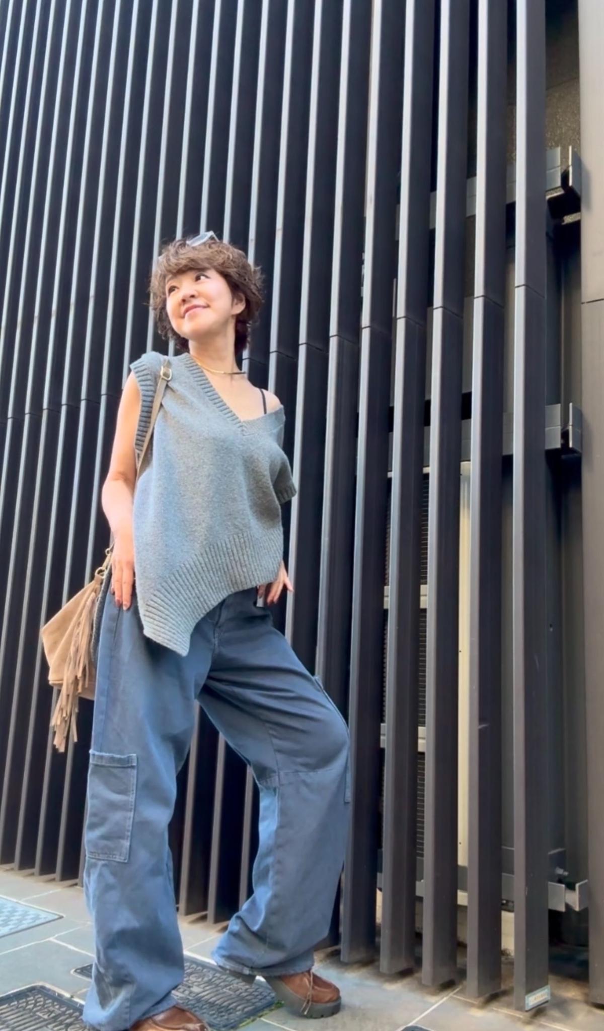 グレーのニットベストとデニムパンツを着用した女性。茶色のバッグを斜め掛けし、短くカットした茶色の髪が特徴的。背景には黒い縦の板が並んだ壁がある。