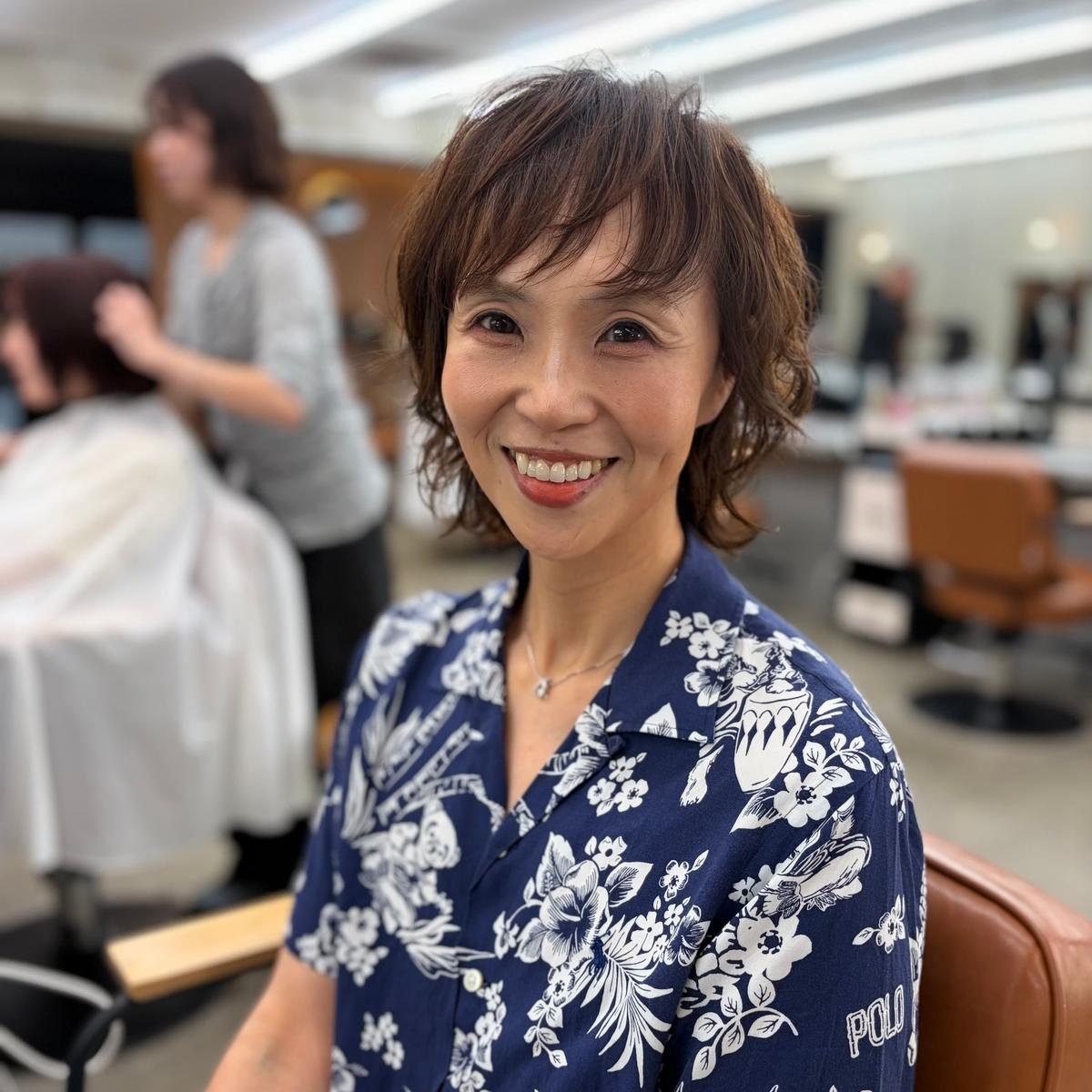 笑顔の女性が、美容室の椅子に座ってこちらを見ている。ダークブルーのfloral柄シャツを着ており、短いダークブラウンの髪に前髪がある。背景には、別の女性が顧客の髪を切っている様子が見える。