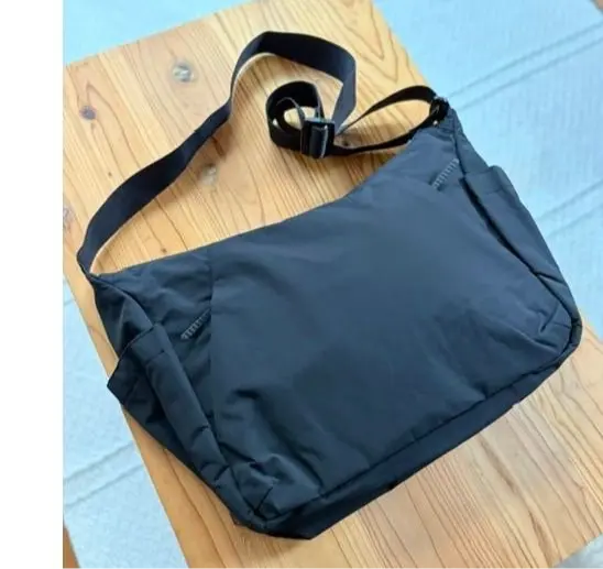 木製のテーブルに置かれた、黒いメッセンジャーバッグ。長めの肩掛けストラップと、前面に複数のポケットがある。シンプルなデザインで、カジュアルな印象。