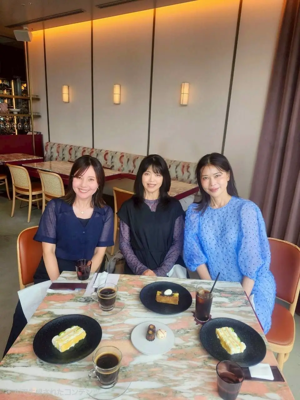 3人の女性がテーブルに座り、デザートとコーヒーを楽しむ様子。テーブルにはサンドイッチ、ケーキ、ドリンクが置かれている。背景には壁の照明とソファが見える。