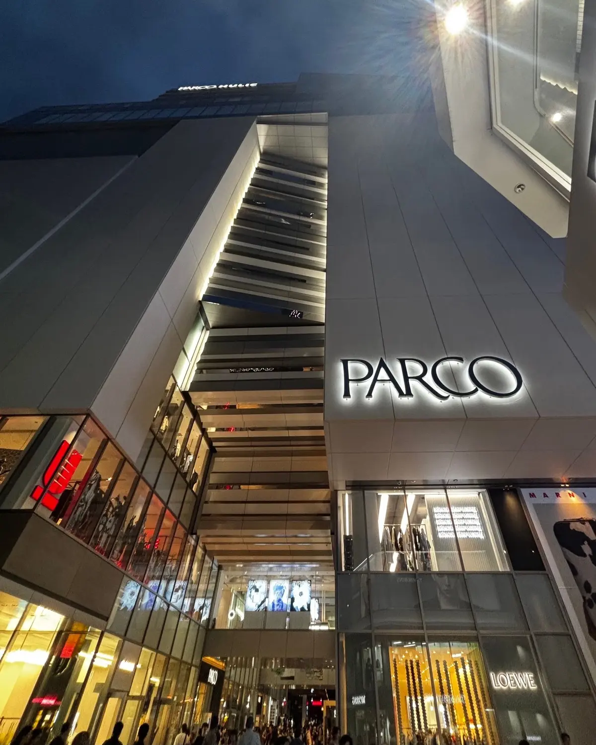 夜の渋谷PARCOの入り口付近。白い屋根が広がり、入り口には大きな「PARCO」のロゴがある。店内は明るく、多くの人が買い物客として歩いている。