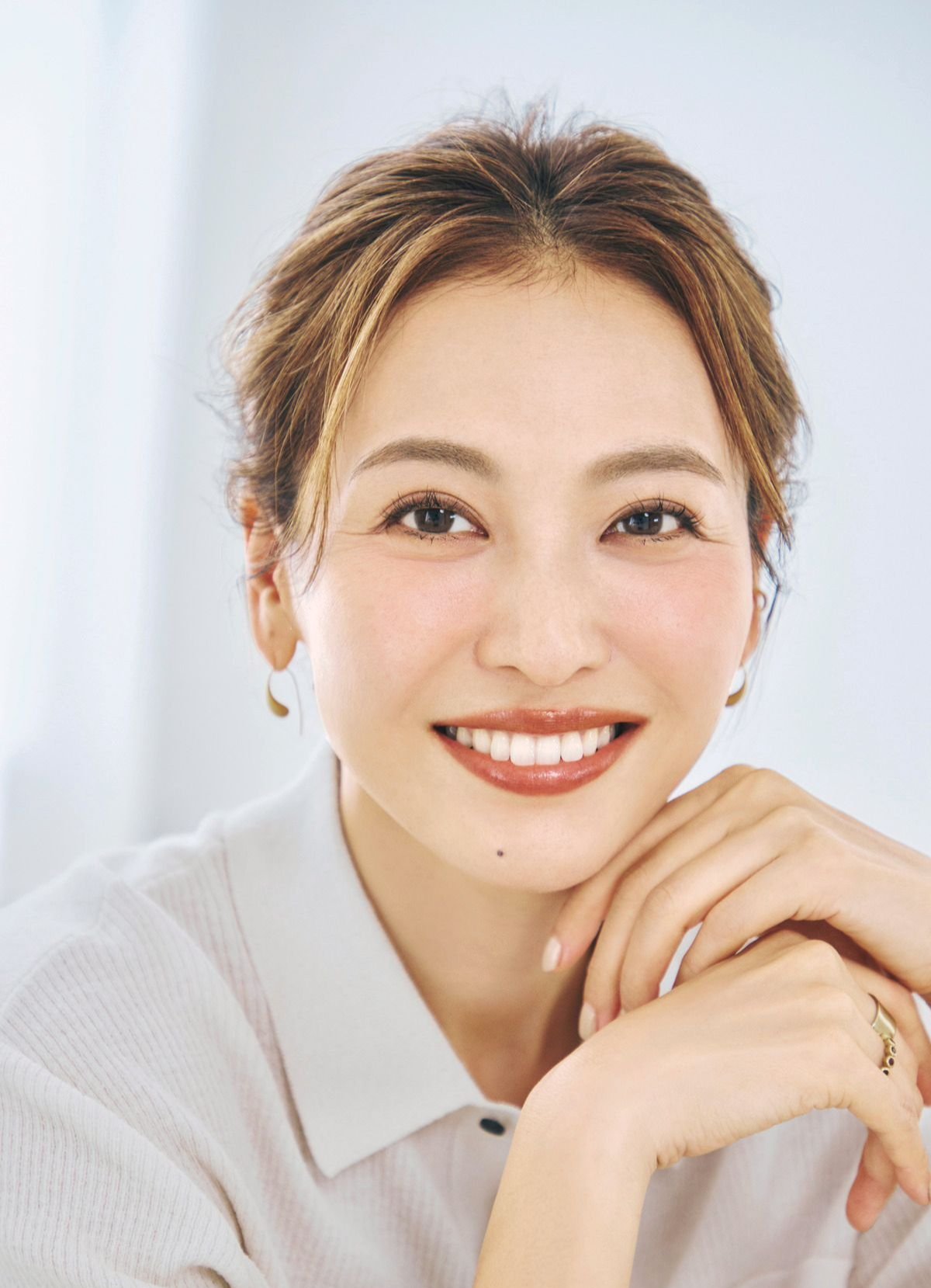 白いシャツを着た女性が微笑むポートレート。茶色の髪をまとめ、ゴールドのイヤリングと腕時計を身に着けている。背景は白。